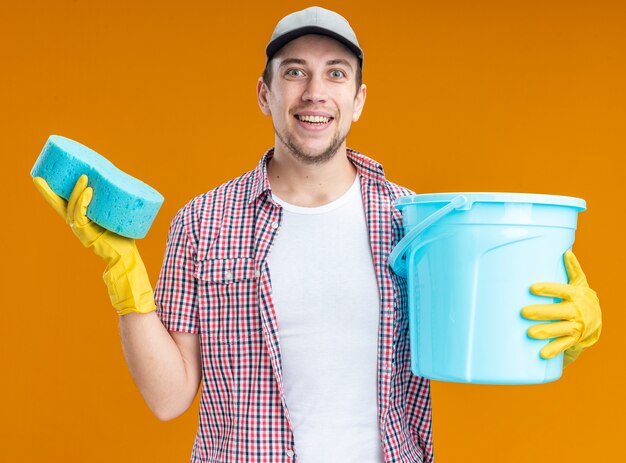 Sonriente joven limpiador con gorra con guantes sosteniendo esponja con balde aislado sobre fondo naranja