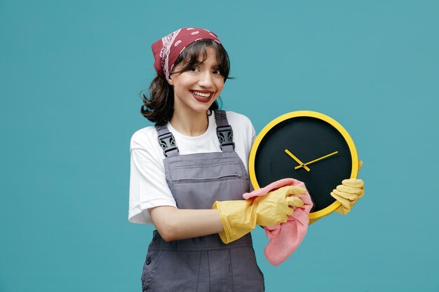 Sonriente joven limpiadora con pañuelo uniforme y guantes de goma sosteniendo el reloj limpiándolo con un plumero mirando la cámara aislada en el fondo azul