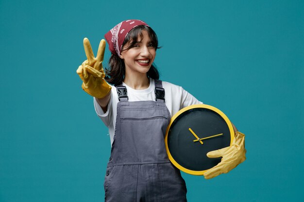 Sonriente joven limpiadora con pañuelo uniforme y guantes de goma sosteniendo el reloj mirando la cámara estirando la mano hacia la cámara mostrando el signo de la paz aislado en el fondo azul