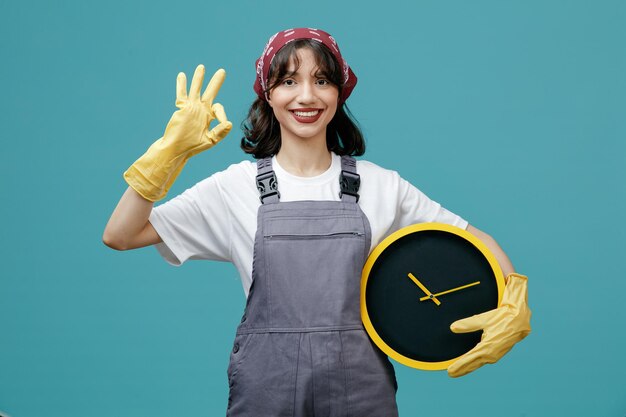Sonriente joven limpiadora con pañuelo uniforme y guantes de goma sosteniendo el reloj mirando a la cámara mostrando el signo de ok aislado en el fondo azul