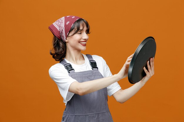 Sonriente joven limpiadora vistiendo uniforme y bandana estirando la bandeja limpiándola con una esponja mirando la bandeja aislada en el fondo naranja