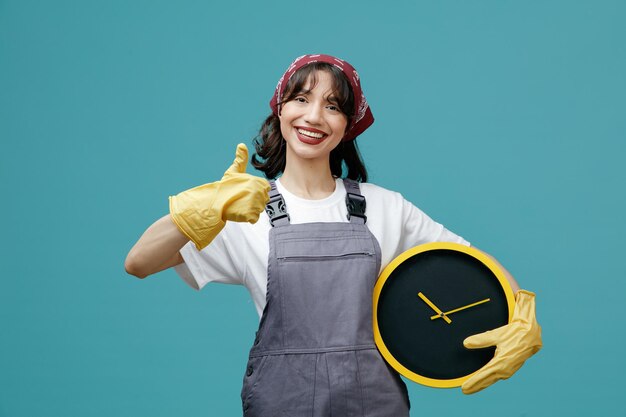 Sonriente joven limpiadora vistiendo uniforme pañuelo y guantes de goma sosteniendo el reloj mirando a la cámara mostrando el pulgar hacia arriba aislado sobre fondo azul.