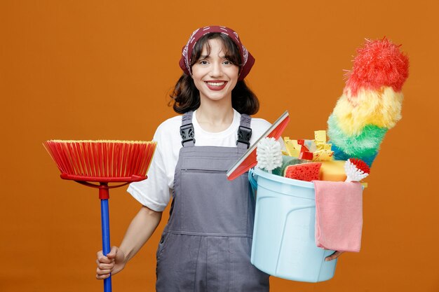 Sonriente joven limpiadora vistiendo uniforme y pañuelo sosteniendo un trapeador y un balde de herramientas de limpieza mirando la cámara aislada en el fondo naranja