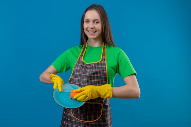 Sonriente joven de limpieza con uniforme en guantes lavando platos sobre fondo azul aislado