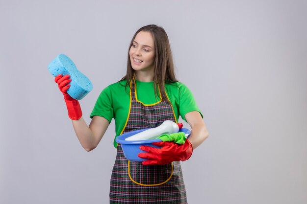 Sonriente joven de limpieza con uniforme en guantes rojos sosteniendo herramientas de limpieza mirando esponja en su mano sobre fondo blanco aislado