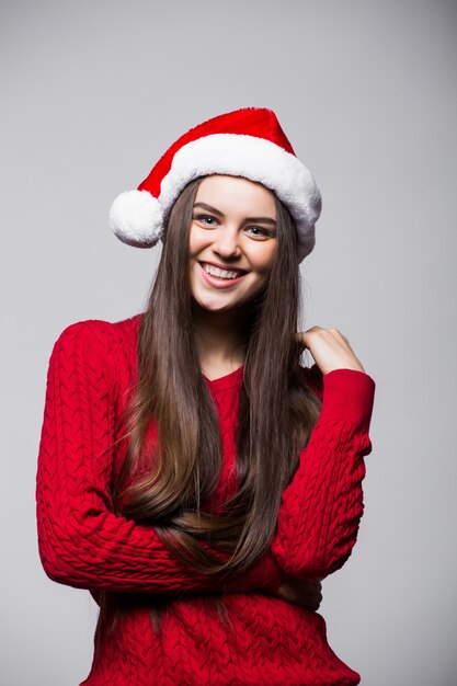 Sonriente joven linda con sombrero de santa claus de pie y mirando hacia arriba sobre la pared gris