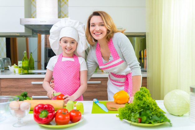 Sonriente joven madre con hija en delantal rosa cocinar verduras en la cocina.
