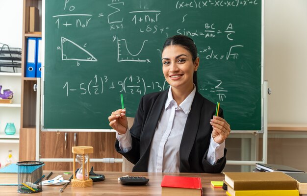 Sonriente joven maestra se sienta a la mesa con útiles escolares sosteniendo un lápiz en el aula