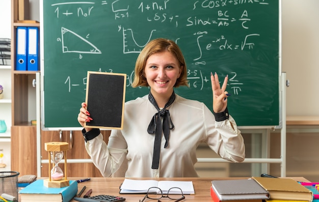 Sonriente joven maestra se sienta a la mesa con útiles escolares sosteniendo una mini pizarra mostrando tres en el aula