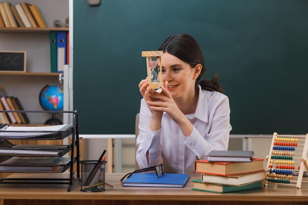 sonriente joven maestra sosteniendo y mirando el reloj de arena sentado en el escritorio con herramientas escolares en el aula