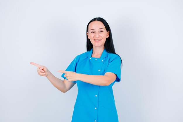 Sonriente joven médico apunta a la izquierda con los dedos índices sobre fondo blanco.