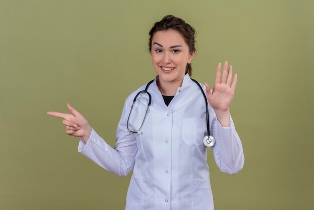 Sonriente joven médico con bata médica con estetoscopio muestra diferentes gestos en la pared verde