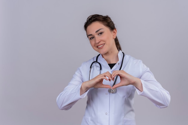Sonriente joven médico con bata médica con estetoscopio muestra gesto de corazón en la pared blanca