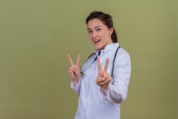 Sonriente joven médico con bata médica con estetoscopio muestra gesto de paz en la pared verde