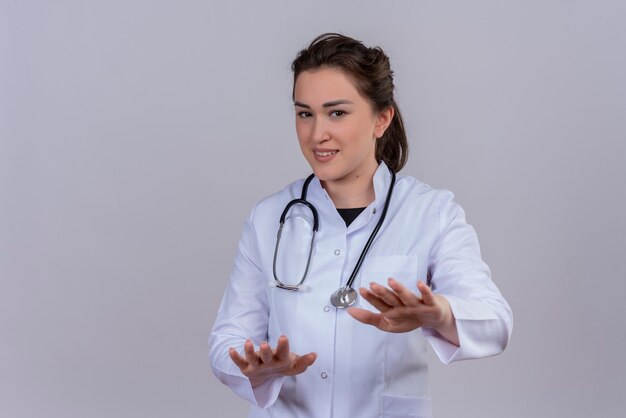 Sonriente joven médico con bata médica con estetoscopio muestra el tamaño en la pared blanca