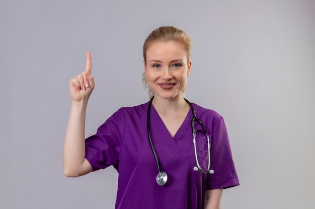 Sonriente joven médico con bata médica púrpura y puntos de estetoscopio hacia arriba en la pared blanca aislada