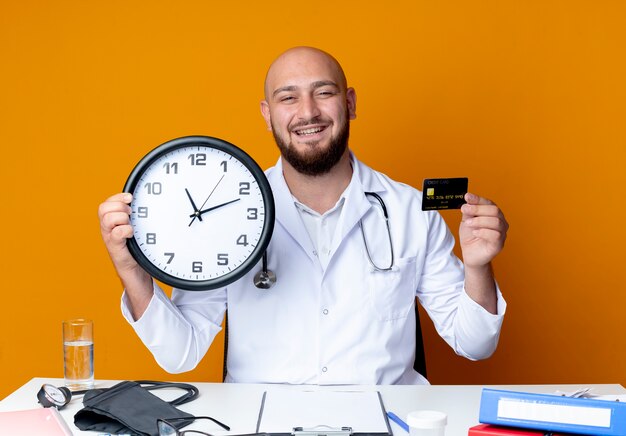 Sonriente joven médico calvo vestido con bata médica y estetoscopio sentado en el escritorio con herramientas médicas con reloj de pared y tarjeta de crédito aislado sobre fondo naranja