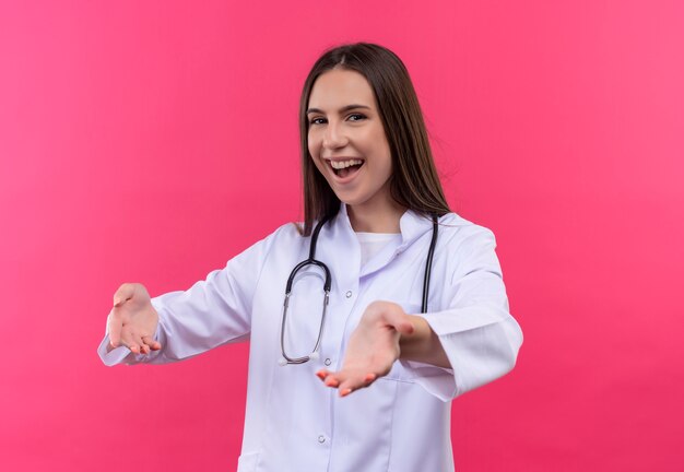 Sonriente joven médico chica vistiendo estetoscopio bata médica fingir sosteniendo algo sobre fondo rosa aislado