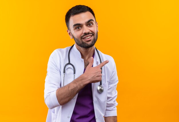 Sonriente joven médico con estetoscopio bata médica apunta al lado con el dedo sobre fondo amarillo aislado