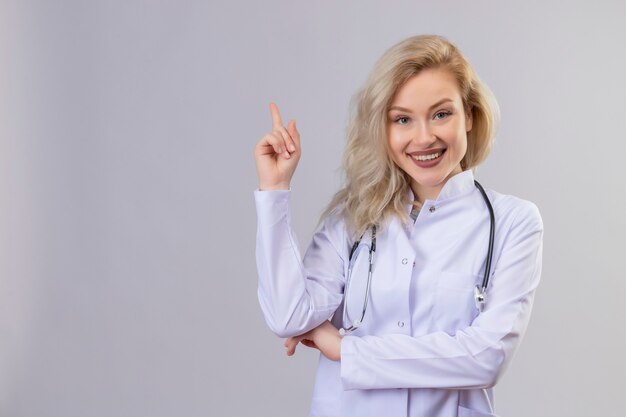 Sonriente joven médico con estetoscopio en bata médica apunta al lado de la pared blanca