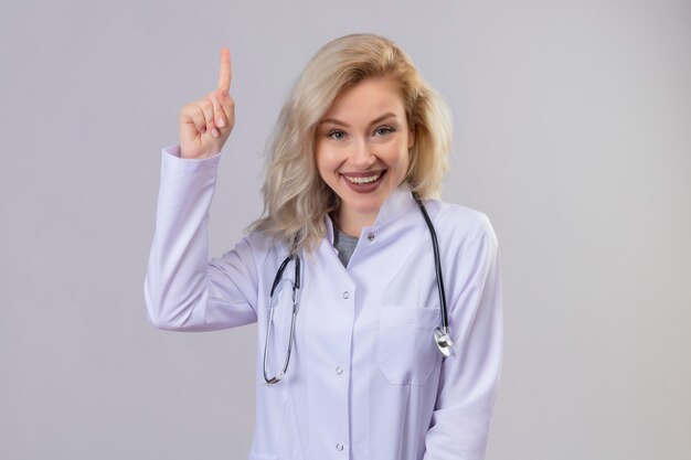 Sonriente joven médico con estetoscopio en bata médica apunta hacia arriba en la pared blanca