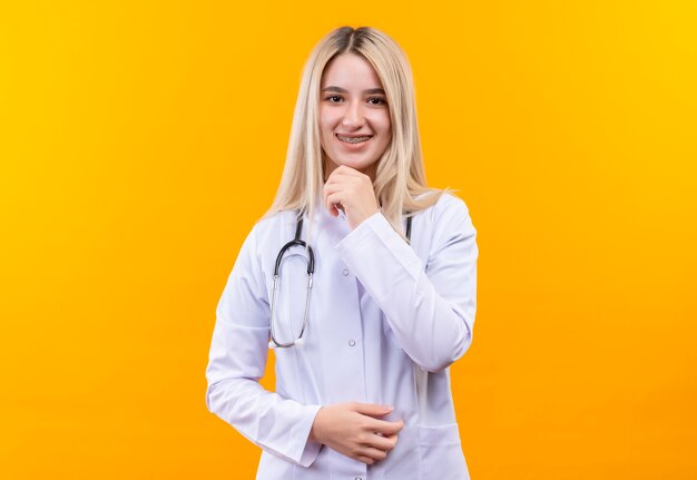 Sonriente joven médico con estetoscopio en bata médica y corsé dental puso su mano en la barbilla sobre fondo amarillo aislado