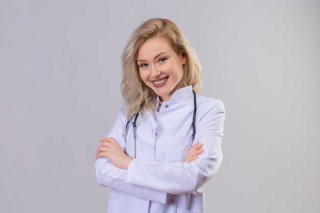 Sonriente joven médico con estetoscopio en bata médica cruzando las manos sobre la pared blanca