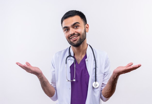 Sonriente joven médico con estetoscopio bata médica extiende las manos sobre fondo blanco aislado