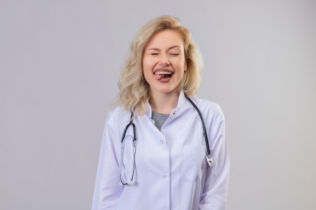 Sonriente joven médico con estetoscopio en bata médica morder la lengua en la pared blanca