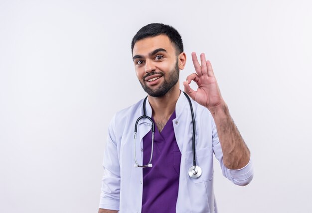 Sonriente joven médico con estetoscopio bata médica mostrando gesto bien sobre fondo blanco aislado