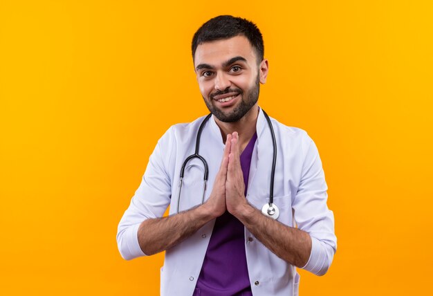 Sonriente joven médico con estetoscopio bata médica mostrando gesto de oración sobre fondo amarillo aislado
