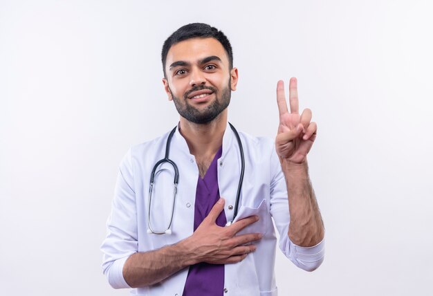 Sonriente joven médico con estetoscopio bata médica mostrando gesto de paz puso su mano sobre el corazón sobre fondo blanco aislado