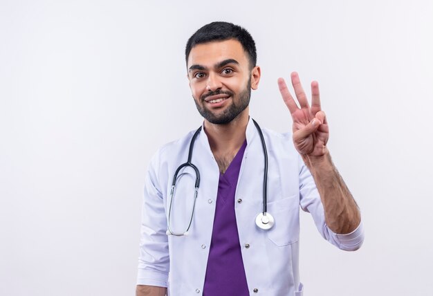 Sonriente joven médico con estetoscopio bata médica mostrando tres sobre fondo blanco aislado