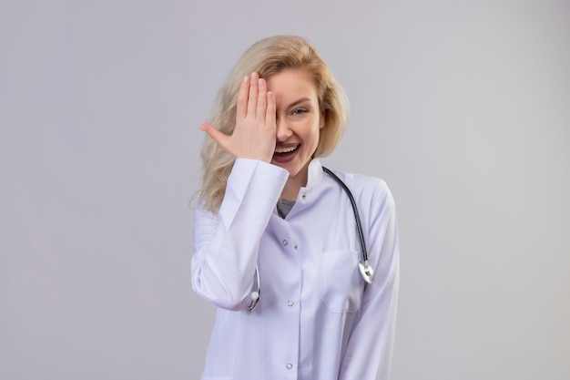 Sonriente joven médico con estetoscopio en bata médica ojo cubierto en pared blanca