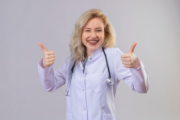 Sonriente joven médico con estetoscopio en bata médica con el pulgar hacia arriba en la pared blanca