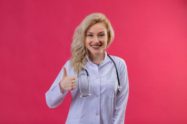 Sonriente joven médico con estetoscopio en bata médica con el pulgar hacia arriba sobre fondo rojo.