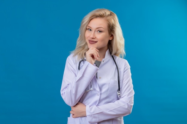 Sonriente joven médico con estetoscopio en bata médica puso su mano debajo de la mandíbula en la pared azul