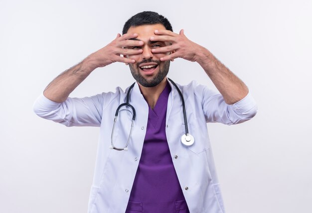 Sonriente joven médico con estetoscopio bata médica puso sus manos sobre los ojos sobre fondo blanco aislado