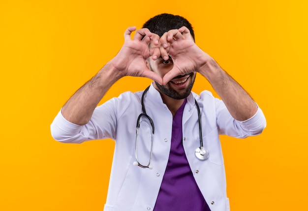 Sonriente joven médico con estetoscopio bata médica que muestra el gesto del corazón sobre fondo amarillo aislado