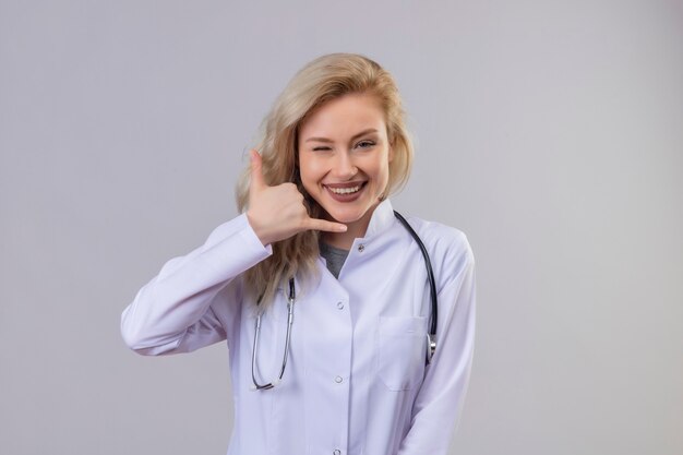 Sonriente joven médico con estetoscopio en bata médica que muestra el gesto de llamada en la pared blanca