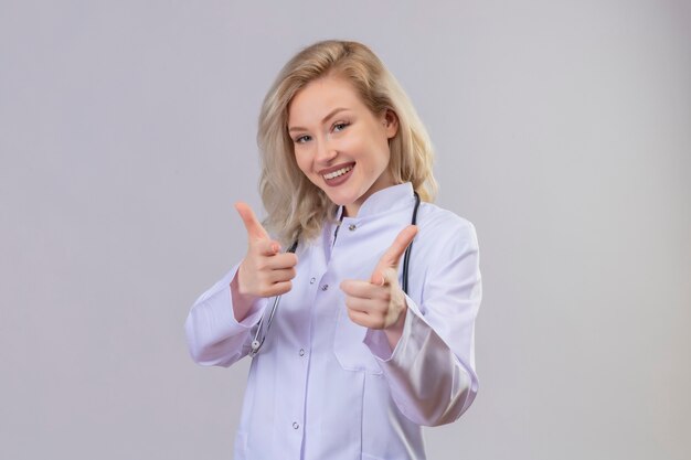 Sonriente joven médico con estetoscopio en bata médica que le muestra gesto en la pared blanca