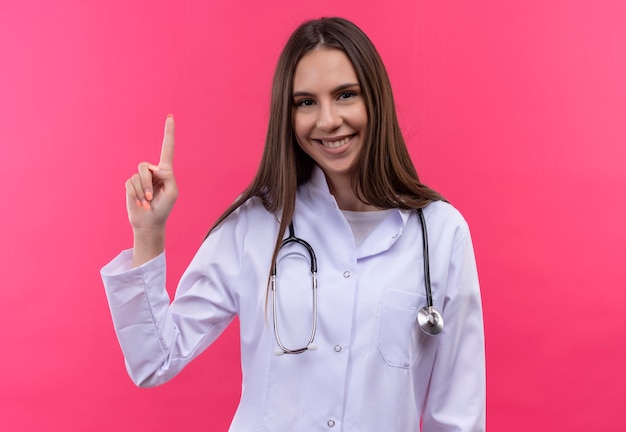 Sonriente joven médico con estetoscopio bata médica señala con el dedo hacia arriba sobre fondo rosa aislado