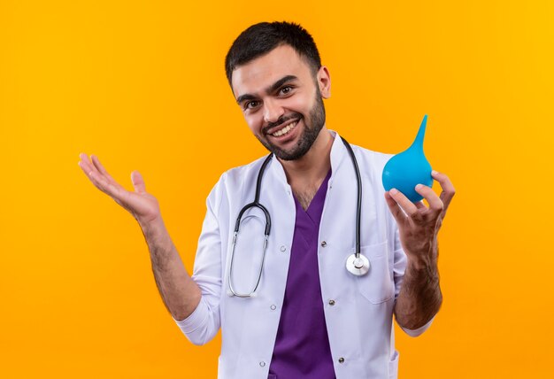Sonriente joven médico con estetoscopio bata médica sosteniendo enema sobre fondo amarillo aislado