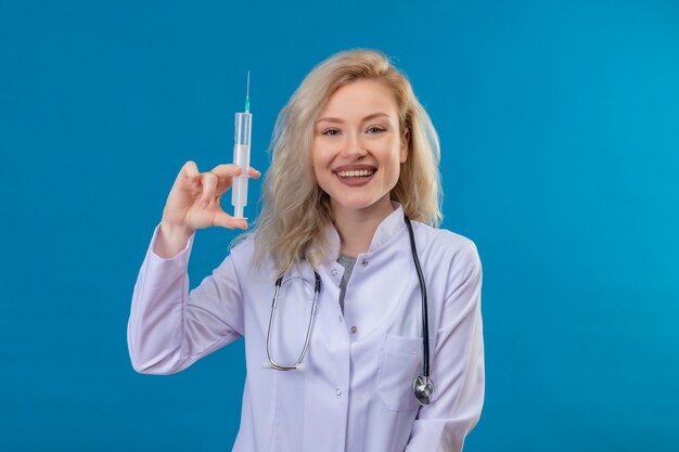 Sonriente joven médico con estetoscopio en bata médica sosteniendo la jeringa en la pared azul