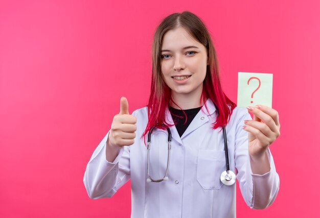 Sonriente joven médico con estetoscopio bata médica sosteniendo papel signo de interrogación con el pulgar hacia arriba sobre fondo rosa aislado