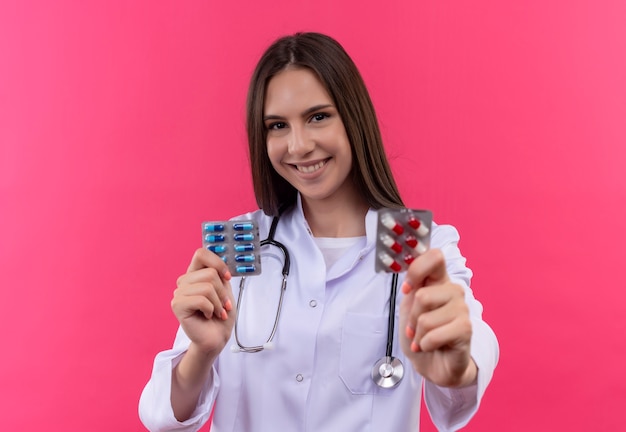 Sonriente joven médico con estetoscopio bata médica sosteniendo pastillas sobre fondo rosa aislado