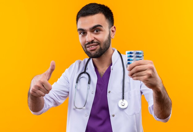 Sonriente joven médico con estetoscopio bata médica sosteniendo píldoras su pulgar hacia arriba sobre fondo amarillo aislado