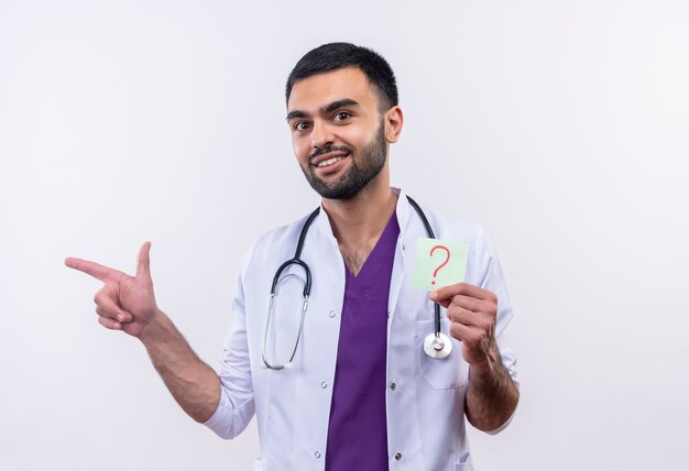 Sonriente joven médico con estetoscopio bata médica sosteniendo puntos de interrogación de papel al lado sobre fondo blanco aislado