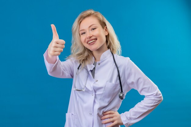 Sonriente joven médico con estetoscopio en bata médica su pulgar hacia arriba en la pared azul
