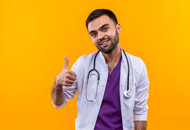 Sonriente joven médico con estetoscopio bata médica su pulgar hacia arriba sobre fondo amarillo aislado